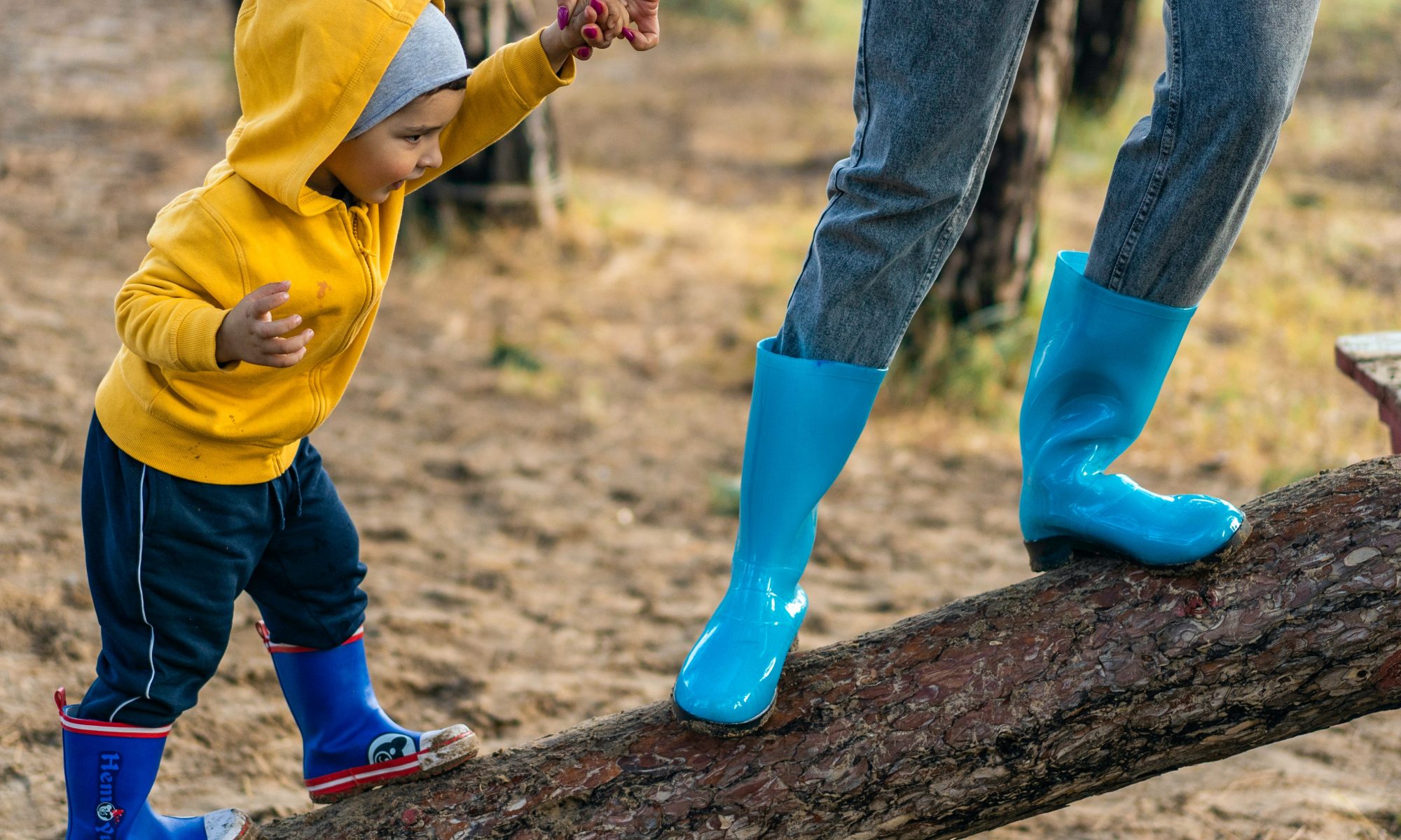 parent-leads-small-child-in-blue-rainboots-on-climb-up-fallen-tree-trunk
