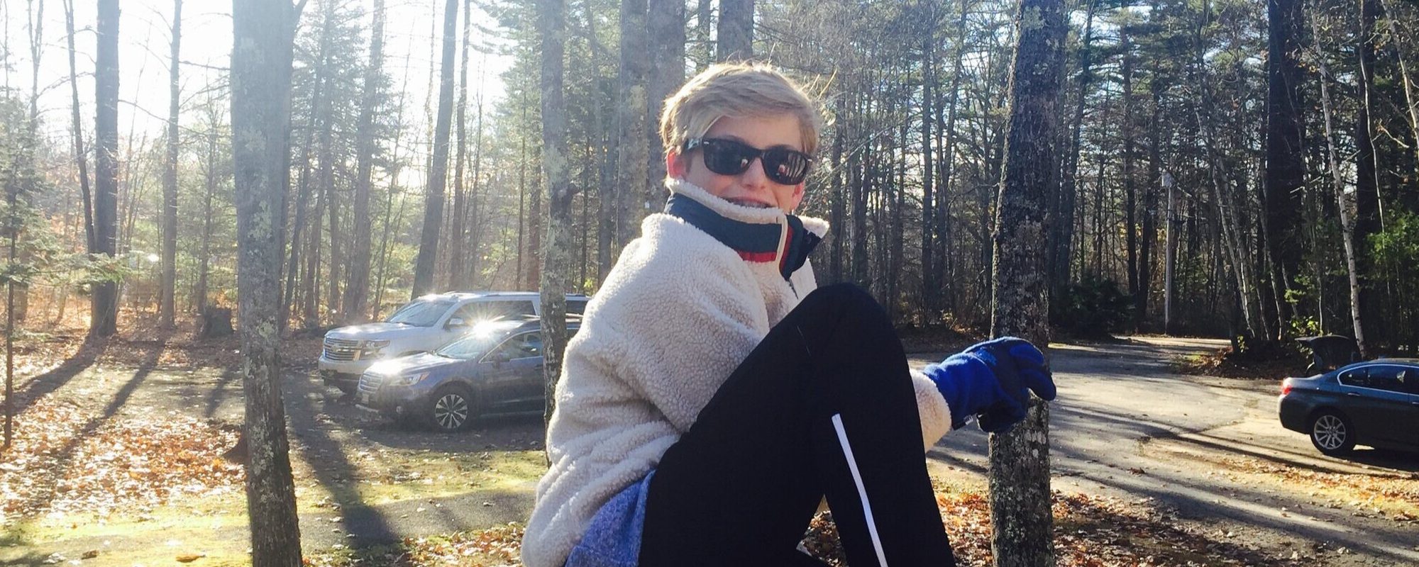 young-boy-wears-sunglasses-smiles-at-camera-on-cold-sunny-day-wooded-background