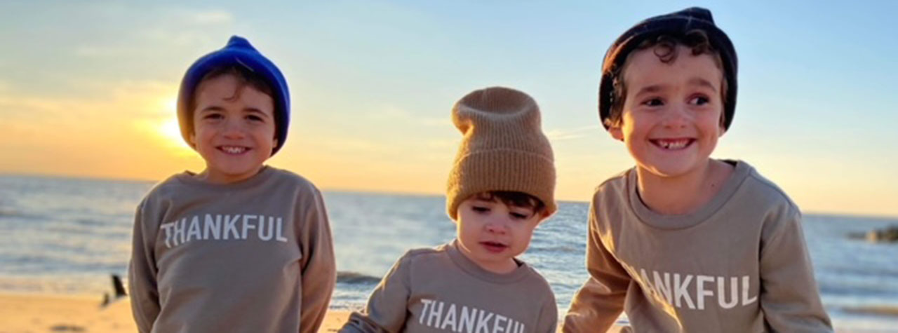 three smiling little brothers at the beach at sunset wear knitted caps and sweatshirts that say THANKFUL