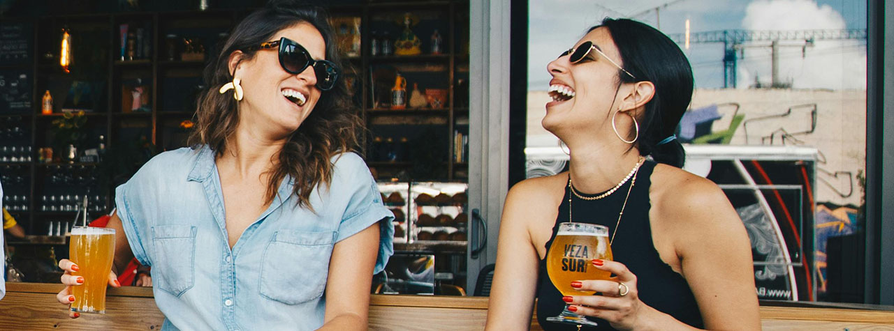 Two women talk, laugh and drink beer sitting on a bench outside a bar. By Elevate on Pexels.