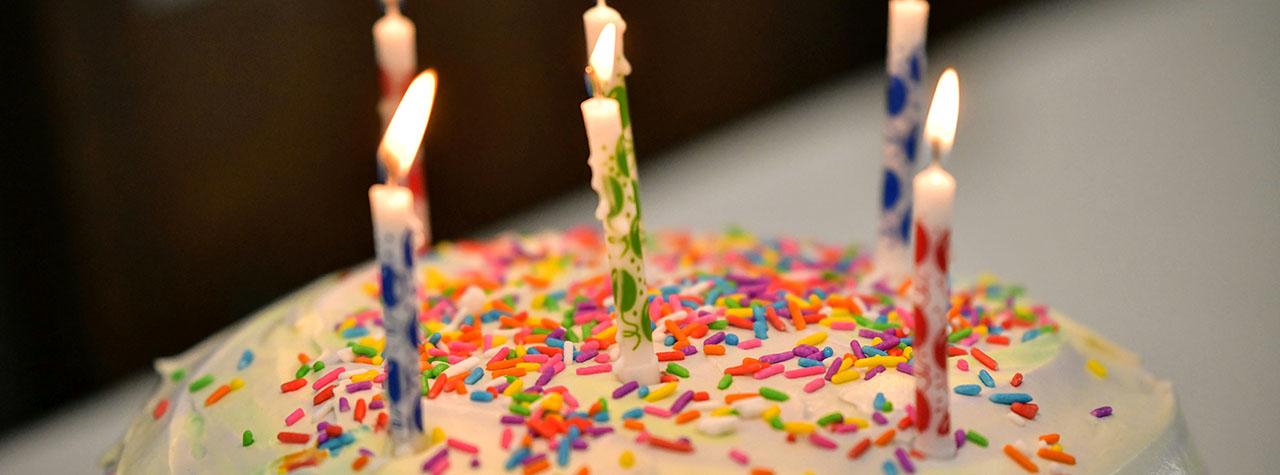 Cake with white frosting, 6 candles, and rainbow sprinkles. Image by Shraga Kopstein on Unsplash.