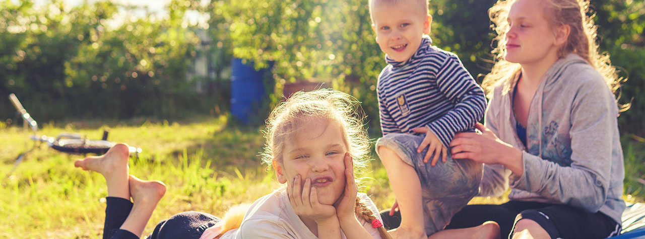 Three fair haired children sit on golden green grass in field surrounded by trees. Image by Vitolda Klein on Unsplash.