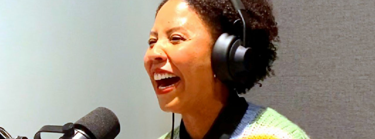 Post author Lindiwe Lewis, wearing a bright yellow sweater and a headset, laughs into a mirophone while podcasting for her new venture Behind the Allergy. Image shared by Lindiwe Lewis.