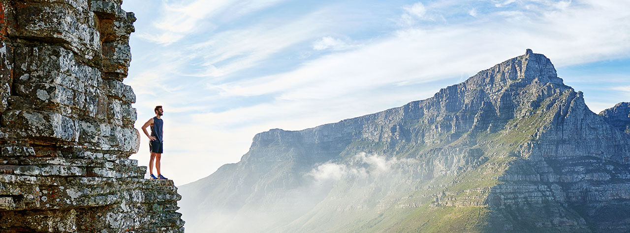 Man in shorts stands on cliff edge overlooking mountain and valley vista. Image by Marius Venter on Pexels.