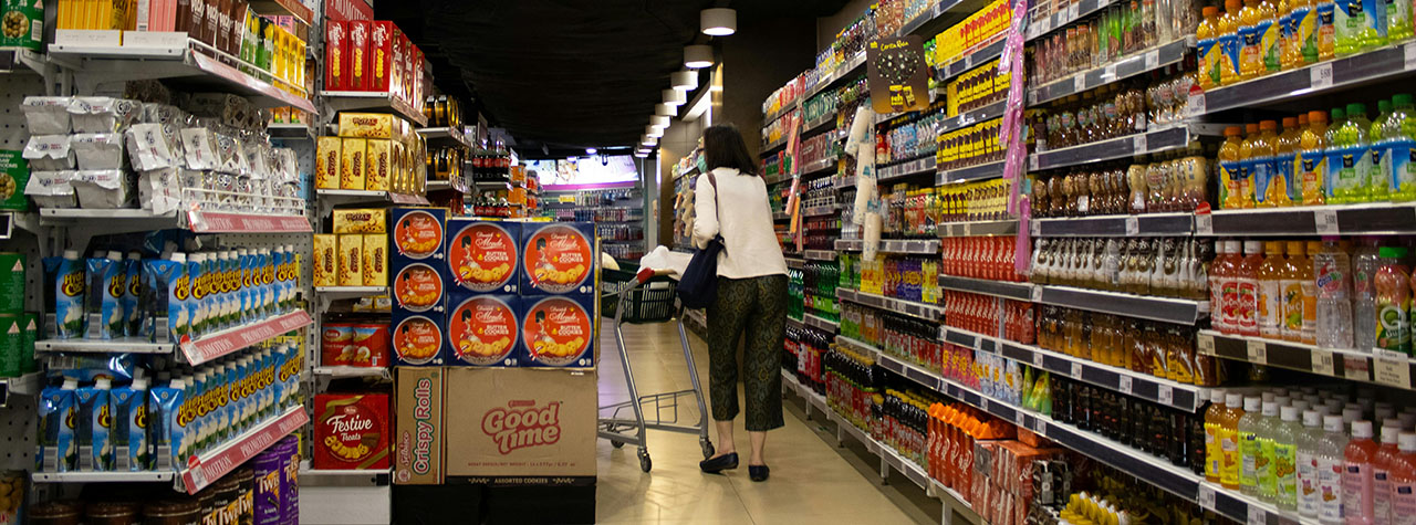 Shopper at grocery store pushes cart down an aisle of food choices. Image by Hobi Industri on Pexels.