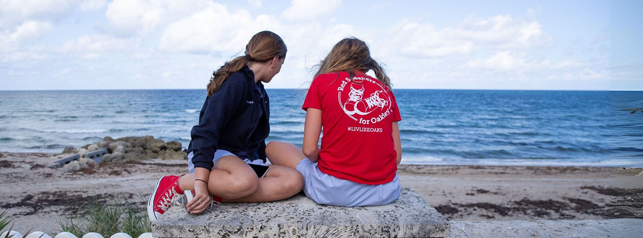 Two girls, one wearing a red 'Red Sneakers for Oakley' t-shirt, the other wearing red sneakers, sit with their backs to the camera. They gaze at a calm, blue ocean. Image from Red Sneakers for Oakley.