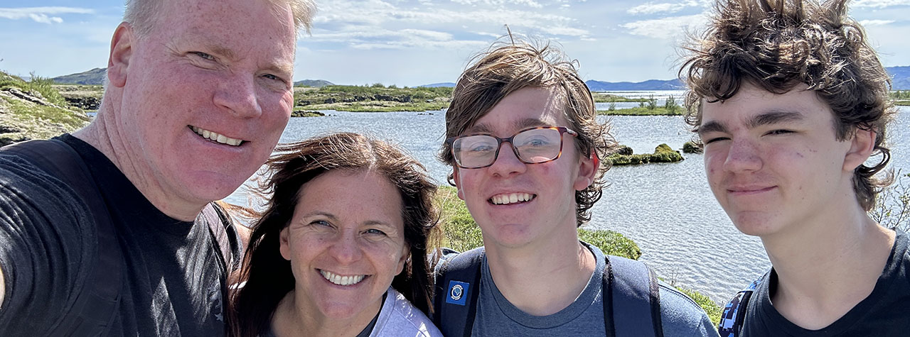 Family of four on vacation stands in front of a body of blue water and rolling hills on a sunny day, smiling at the camera. Image courtesy of The Allergy Aspect.