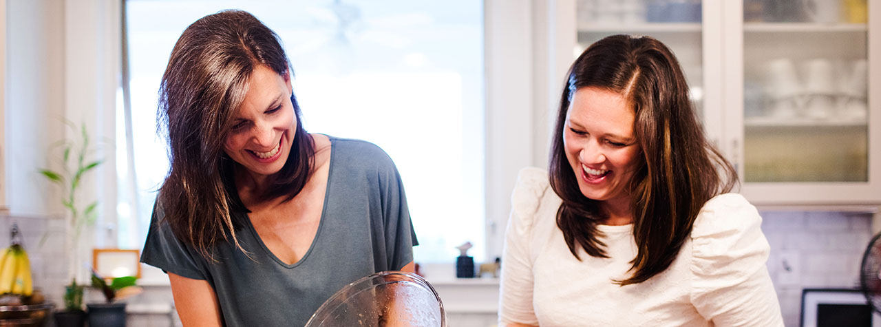 Two women with dark hair laugh together as they prepare food in the kitchen. Image courtesy of Two Alpha Gals.