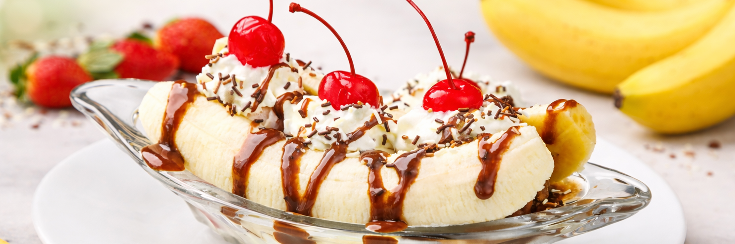 A banana split in a clear glass dish has bananas sliced in half with three scoops of vanilla ice cream topped with whipped cream, three maraschino cherries, chocolate sauce, and sprinkles. Image by Eiliv Aceron on Pexels.
