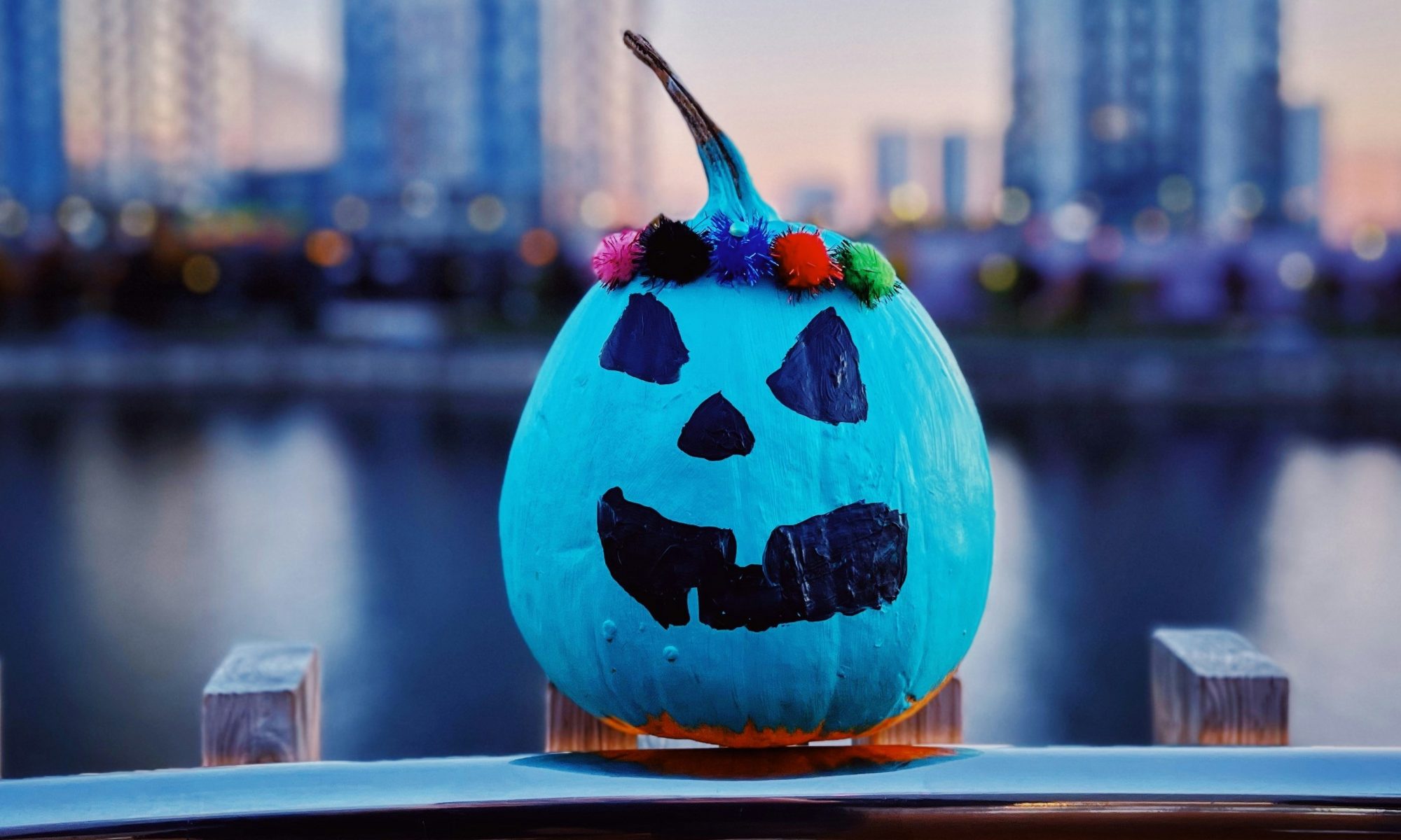 Teal pumpkin with painted face sits on a fence with a city backdrop behind wearing a multi-color crown. Image by Igor Karimov on Unsplash.